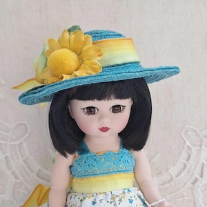 May include: A doll with black hair and brown eyes wearing a blue hat with a yellow sunflower accent. The doll is dressed in a blue and yellow top and a floral print dress, creating a cheerful, summery look.