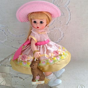 May include: A vintage doll with blonde hair, blue eyes, and red lips, wearing a pink dress, hat, and white shoes. The doll is holding a brown bunny ornament with a pink flower. The dress has floral and yellow accents.