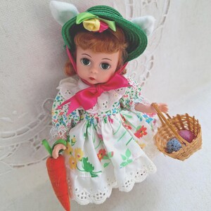 May include: A vintage doll with red hair, wearing a green hat with floral accents and bunny ears. The doll is dressed in a floral print dress with a pink bow and holds a carrot and a basket with colorful eggs. The doll is approximately 10 inches tall.
