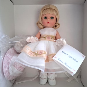 May include: A Madame Alexander Easter Splendor doll in a white dress with floral accents. The doll has blonde hair, green eyes, and is wearing white shoes. The doll is in its original packaging.