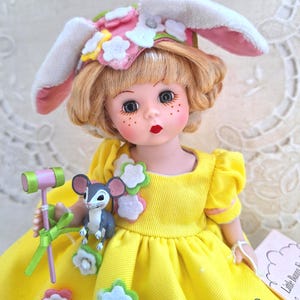 May include: A vintage doll with blonde hair, wearing a yellow dress and a bunny ear hat with floral accents. The doll holds a miniature hammer and a gray mouse figurine. The doll has rosy cheeks and red lips.