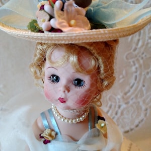 May include: A porcelain doll wearing a blue dress and a straw hat with a bird and flowers. The doll has blonde hair, blue eyes, and a delicate face.