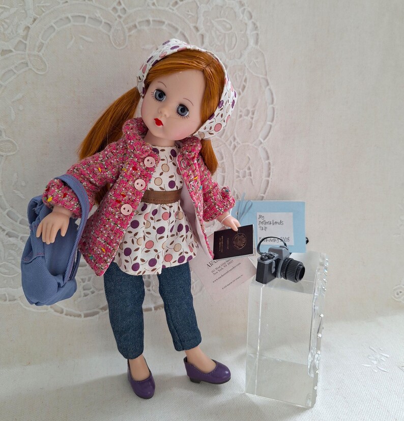 May include: A red-haired doll wearing a pink and white patterned jacket, a white and blue patterned headband, blue jeans, and purple shoes. The doll is holding a blue backpack and a passport. The doll is standing on a white surface with a small camera and a note that says "My Netherlands Trip 2023".