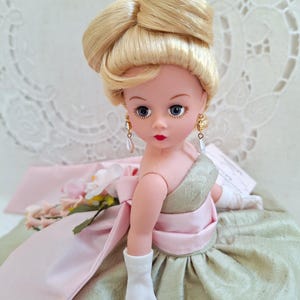 May include: A vintage doll with blonde hair styled in an updo. The doll wears a green and pink dress, gold earrings with pearl drops, and white gloves. A pink sash and floral accents adorn the dress. The doll has blue eyes and red lipstick.