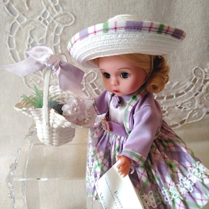 May include: A porcelain doll wearing a lavender and green plaid dress with a white hat. The doll is holding a white wicker basket with lavender flowers and green leaves. The doll is sitting on a clear acrylic display stand. The doll is labeled "Easter" and "Alexander".