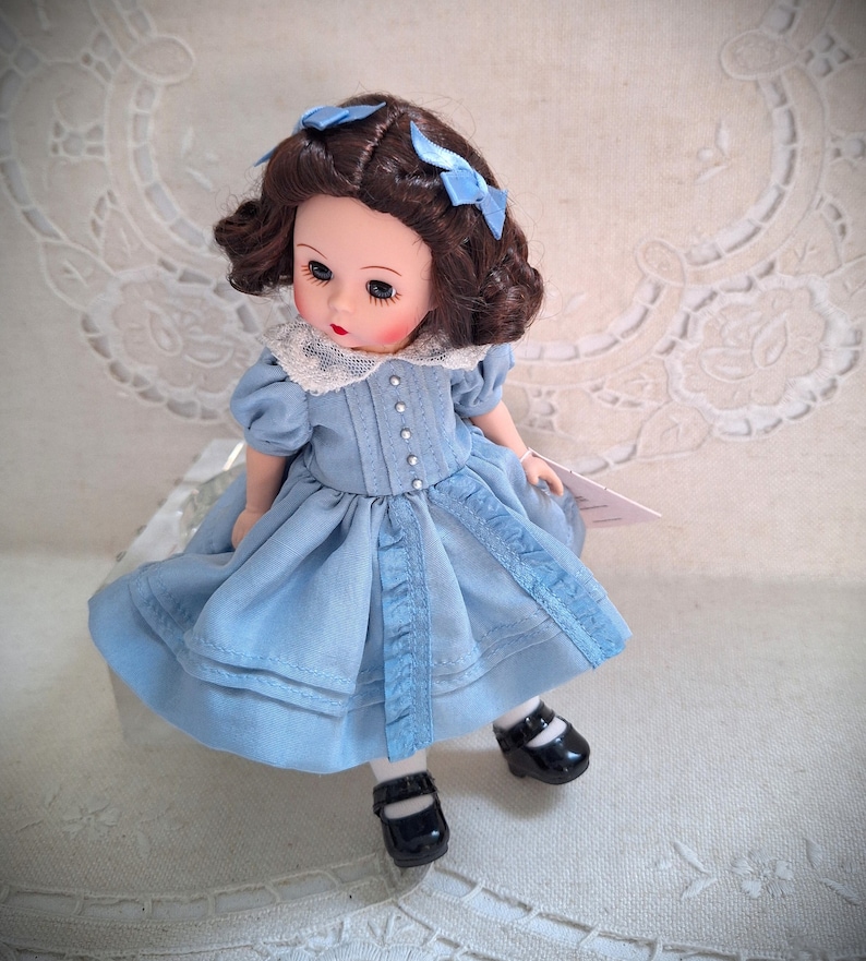Puede incluir: Una mu&ntilde;eca de porcelana con cabello casta&ntilde;o y ojos azules, vestida con un vestido azul claro con ribete de encaje blanco y zapatos negros. La mu&ntilde;eca est&aacute; sentada sobre una superficie blanca.