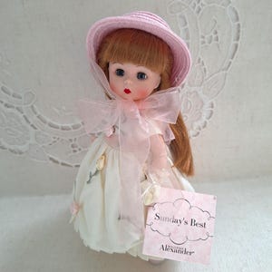 May include: A Madame Alexander doll wearing a pink hat and a white dress with floral accents. The doll has red hair, blue eyes, and a pink bow. A tag reads "Sunday's Best".