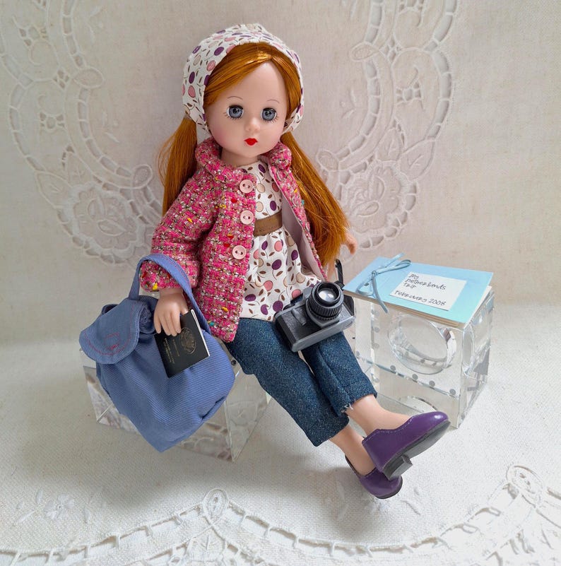 May include: A red-haired doll wearing a pink and white patterned jacket, a blue denim skirt, and purple shoes. The doll is holding a camera and a passport. The doll is sitting on a white surface with a blue backpack and a small white box with the text "My Adventures Trip" and "February 2008".