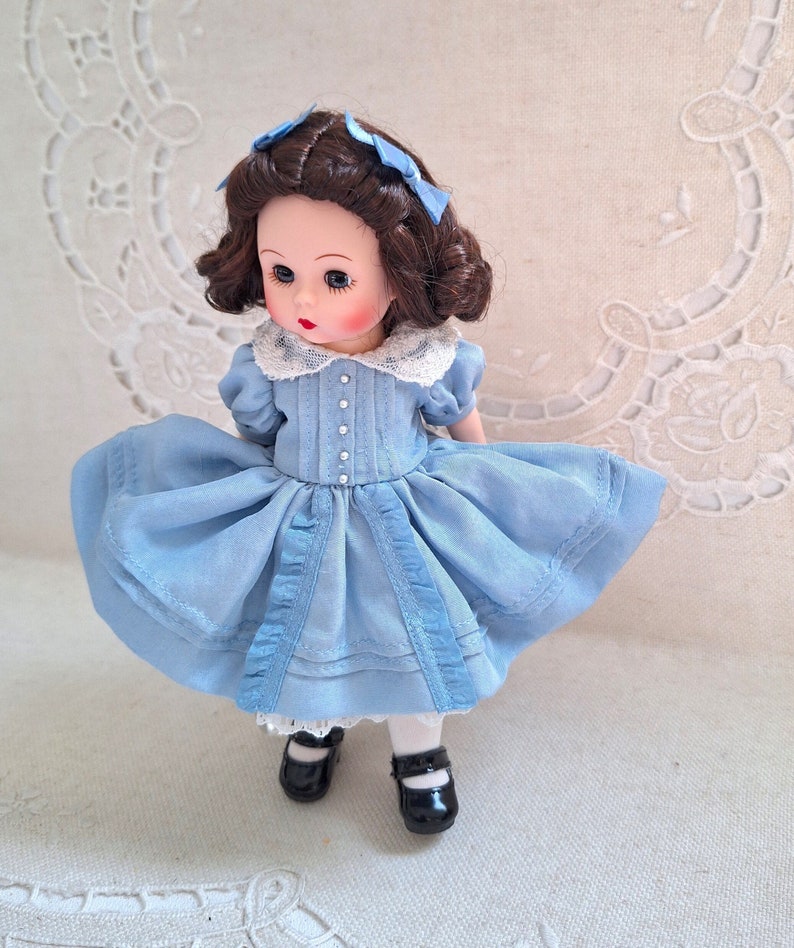 Puede incluir: Una mu&ntilde;eca de porcelana con un vestido azul claro con ribetes de encaje blanco y detalles de cinta azul. La mu&ntilde;eca tiene el pelo casta&ntilde;o con lazos azules y lleva zapatos negros.
