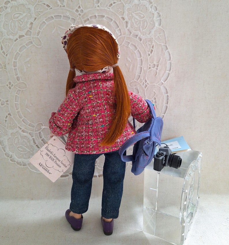 May include: A red-haired doll wearing a pink and white patterned jacket, blue jeans, and a white and brown patterned hat. The doll is carrying a blue backpack and a camera. The doll is standing in front of a white background with a lace pattern.