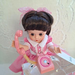 May include: A vintage doll with dark hair and a pink gingham dress holds a pink rotary phone. The doll has a white collar, a pink bow, and freckles. The phone is on a clear stand.