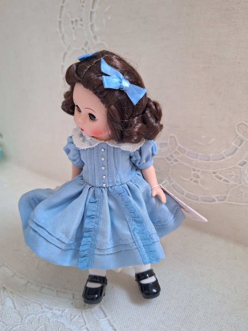 Puede incluir: Una mu&ntilde;eca de porcelana con cabello casta&ntilde;o que lleva un vestido azul claro con ribete de encaje blanco y zapatos negros. La mu&ntilde;eca tiene ojos azules y lleva dos lazos azules en el pelo.
