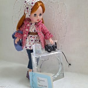 May include: A doll wearing a pink and white patterned jacket, a white and purple patterned dress, and a blue and white patterned headscarf. The doll is holding a small black camera and a passport. The doll is standing on a clear acrylic block with a blue and white card that says "My Netherlands Trip - February 2008".