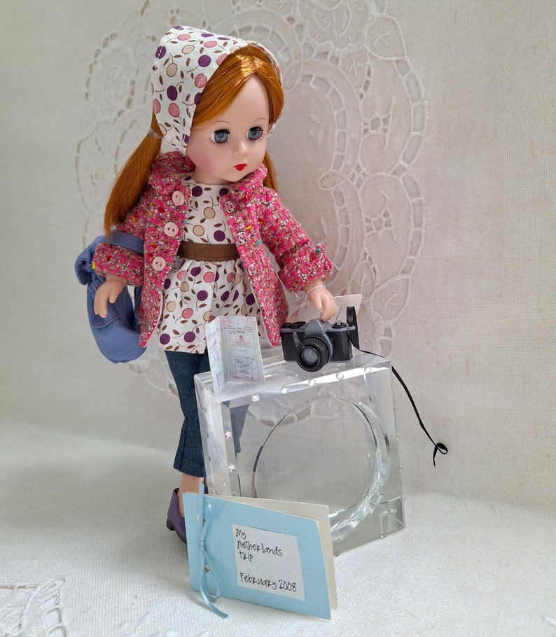 May include: A doll wearing a pink and white patterned jacket, a white and purple patterned dress, and a blue and white patterned headscarf. The doll is holding a small black camera and a passport. The doll is standing on a clear acrylic block with a blue and white card that says "My Netherlands Trip - February 2008".