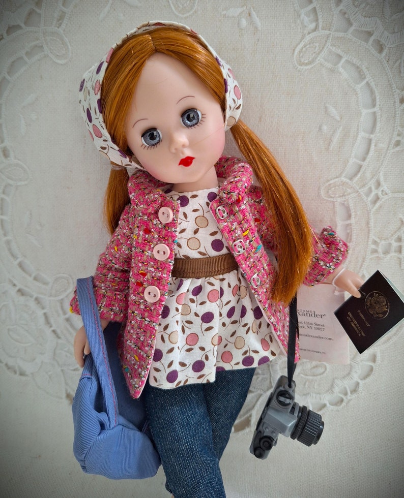 May include: A red-haired doll wearing a pink and white polka dot dress, a brown belt, a pink jacket, a blue bag, and a gray camera. The doll is holding a passport with the words "United States of America" on it.