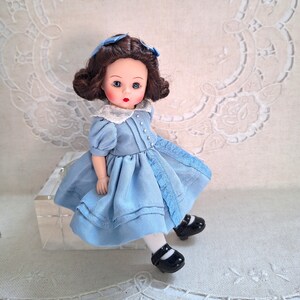 Puede incluir: Una mu&ntilde;eca de porcelana con cabello casta&ntilde;o, ojos azules y un vestido azul claro. Lleva zapatos negros y est&aacute; sentada sobre una superficie blanca.