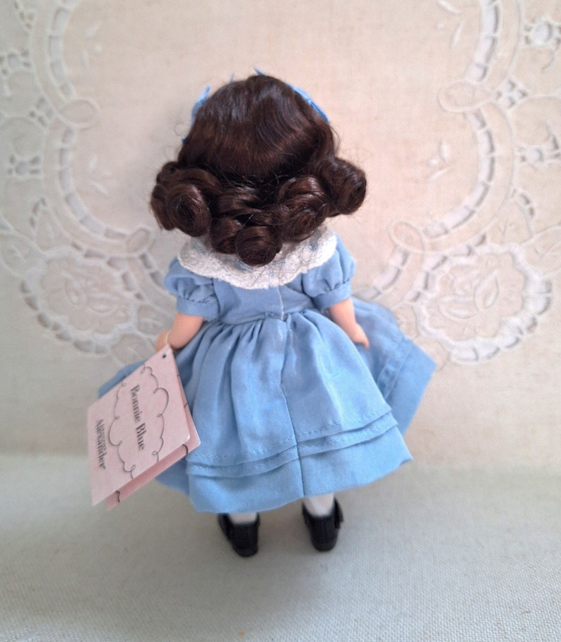Puede incluir: Una peque&ntilde;a mu&ntilde;eca de porcelana que lleva un vestido azul con un cuello de encaje blanco. La mu&ntilde;eca tiene el pelo casta&ntilde;o recogido en dos mo&ntilde;os y sostiene una etiqueta que dice "Jennie Blue" y "Alexander".
