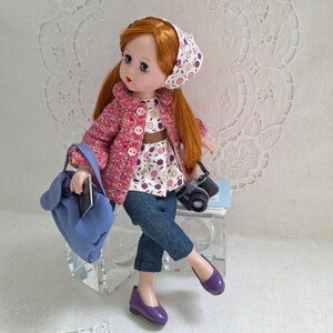 May include: A doll with red hair wearing a pink jacket, a white and purple patterned blouse, blue jeans, and purple shoes. The doll is holding a blue bag and a camera.