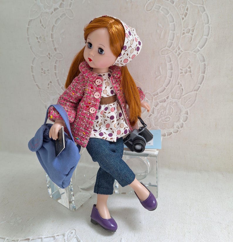 May include: A doll with red hair wearing a pink jacket, a white and purple patterned blouse, blue jeans, and purple shoes. The doll is holding a blue bag and a camera.