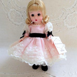 Könnte beinhalten: Vintage Puppe mit blonden Locken, in einem rosa Kleid mit weißer Spitze und einem schwarzen Band gekleidet. Die Puppe trägt schwarze Schuhe und ein Etikett mit der Aufschrift "60th Anniversary Bride" und "Madame Alexander".