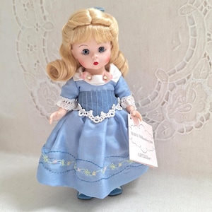 May include: A vintage doll with blonde hair styled in curls, wearing a light blue dress with white lace trim and floral embroidery. The doll has blue eyes and is holding a tag. The doll is a collectible item.