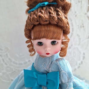 May include: A vintage doll with auburn hair styled in curls and bangs, adorned with a blue bow. The doll has green eyes, rosy cheeks, and red lips. She wears a light blue dress with a large bow at the front.
