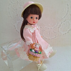 May include: A pink porcelain doll with brown hair wearing a pink dress with white lace trim and a straw hat. The doll is holding a basket filled with Easter eggs and a small bunny.