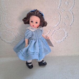 Puede incluir: Una mu&ntilde;eca de porcelana con cabello casta&ntilde;o, ojos azules y un vestido azul. La mu&ntilde;eca lleva zapatos negros y tiene un cuello blanco con detalles de perlas.