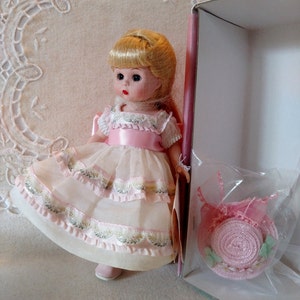 May include: A vintage doll with blonde hair, wearing a pink and white dress with a matching hat. The doll's dress has layered ruffles and decorative trim. The hat is in a clear plastic bag.