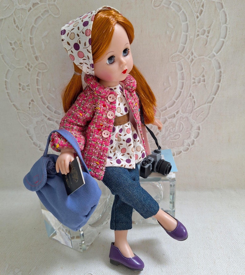 May include: A doll with red hair wearing a pink and white patterned jacket, blue jeans, and purple shoes. She is holding a blue bag and a camera.