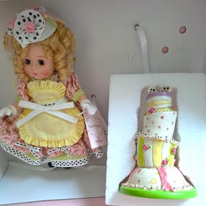 May include: A vintage doll with blonde curly hair, wearing a pink and yellow dress with a matching hat. Next to the doll is a decorative, three-tiered cake ornament with pink and yellow accents, and a green base.