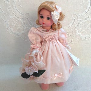 May include: A vintage doll with blonde hair styled with a white bow. The doll wears a pink dress with a floral embroidered collar and carries a matching pink floral purse. The doll's dress has pink bows on the skirt.