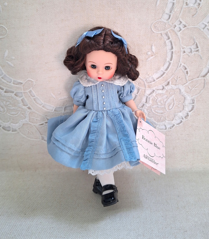 Puede incluir: Una mu&ntilde;eca de porcelana con un vestido azul, un cuello de encaje blanco y medias blancas. La mu&ntilde;eca tiene el pelo casta&ntilde;o y los ojos azules. La mu&ntilde;eca sostiene una etiqueta rosa que dice "Bonnie Blue" y "Alexander Doll Company".
