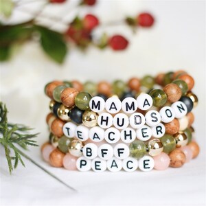 May include: Stack of five beaded bracelets with different words spelled out on each. The words are "Mama", "Hudson", "Teacher", "BFF", and "Peace". The beads are a mix of wood, green, and pink.