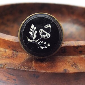 May include: A black and white cameo with a floral design set in a brass ring. The cameo is sitting on a wooden surface.