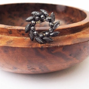 May include: A silver ring with a wreath design made of small, pointed leaves. The ring is sitting on a wooden surface.