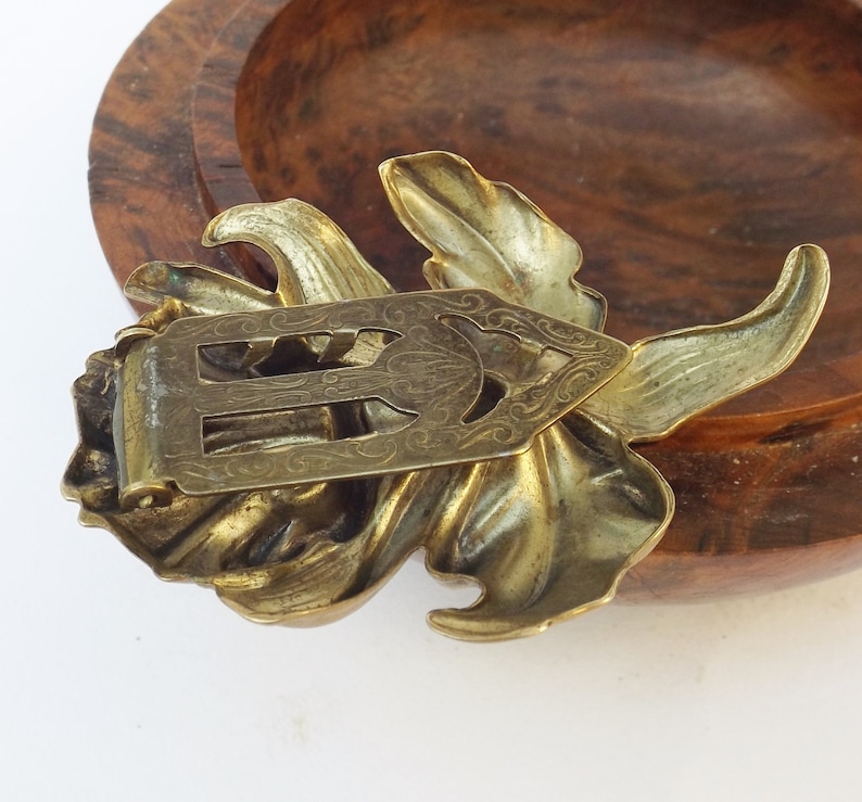 May include: A gold-toned metal flower brooch with an intricate design. The brooch has a floral shape with a detailed center and a clasp on the back.