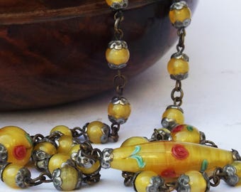 May include: A yellow glass beaded necklace with a large yellow glass pendant featuring painted red and green flowers. The necklace is strung on a silver chain.