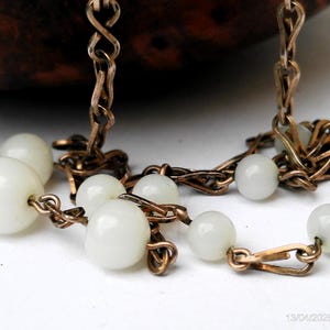 May include: A close-up shot of a vintage necklace. It features off-white, round beads strung on a gold-toned chain with an intricate design. The necklace is set against a white background, with a portion of a dark brown object visible.