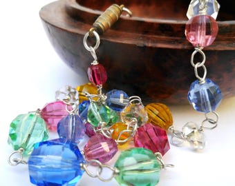 May include: A multi-coloured beaded necklace featuring faceted, round beads in blue, pink, green, yellow, and clear. The beads are strung on a silver-coloured chain, with a brass clasp. The necklace is near a wooden bowl.