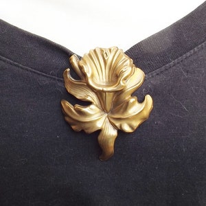 May include: A gold-toned metal flower brooch with a detailed design. The flower has a large, open center and petals that are slightly curled.