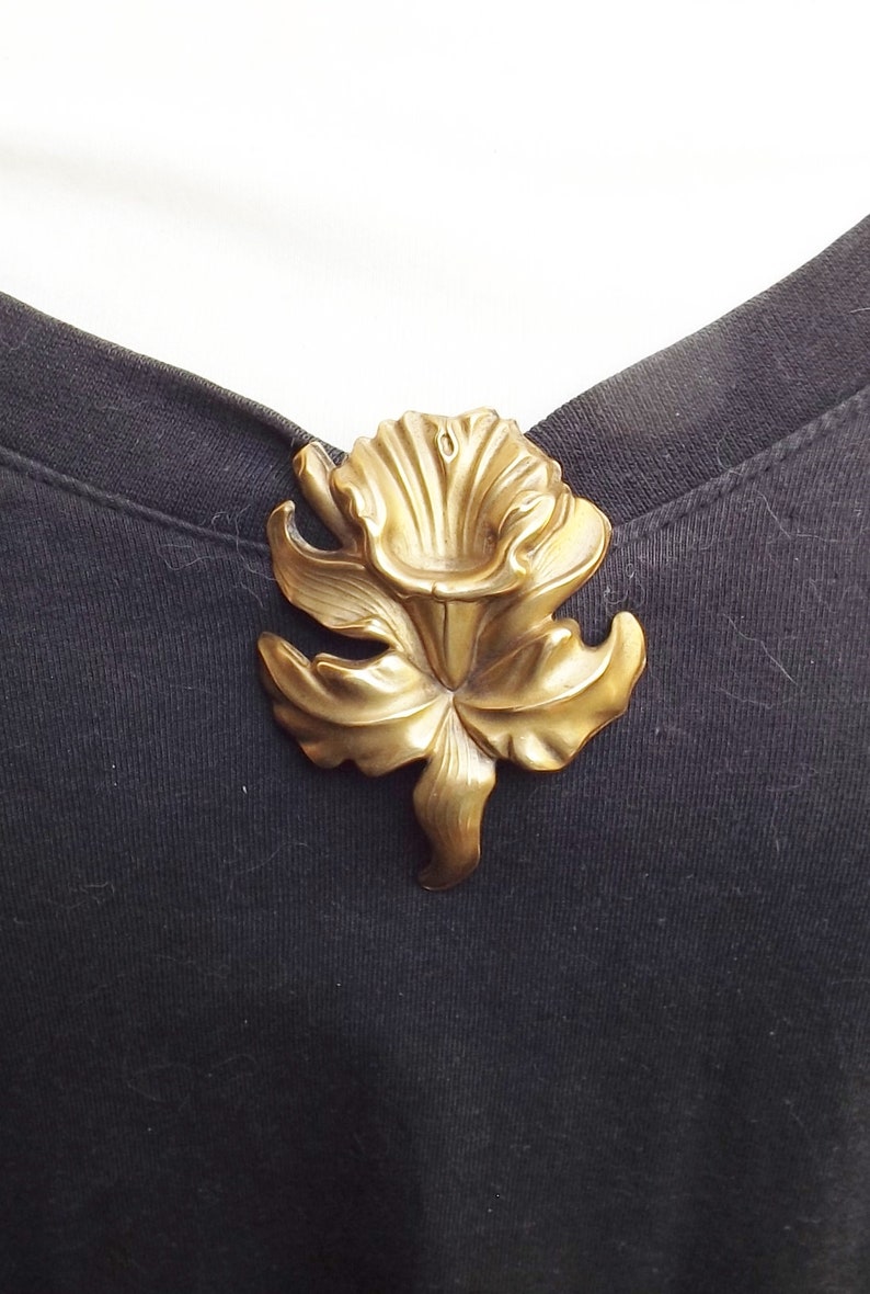 May include: A gold-toned metal flower brooch with a detailed design. The flower has a large, open center and petals that are slightly curled.