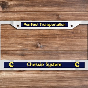May include: A silver-colored license plate frame with the text "PurrFect Transportation" at the top and "Chessie System" at the bottom. The text is yellow on a navy blue background. The frame is on a wooden background.