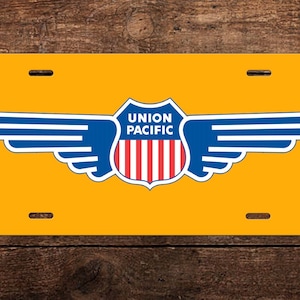 May include: A yellow metal license plate with a blue and red Union Pacific logo featuring a shield with wings. The shield has the words "Union Pacific" in white text.