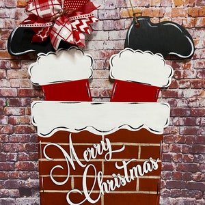 May include: A wooden Christmas door hanger with a red and white Santa Claus boot sticking out of a brick chimney. The chimney has a white snow cap and the words "Merry Christmas" written in white cursive on the front.