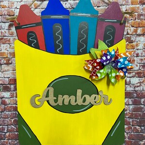 May include: A wooden crayon box-shaped sign with the name "Amber" in gold lettering. The sign features four crayon tops in red, blue, teal, and purple. A decorative bow with various patterns and colors is attached to the front. The sign is designed to hang.