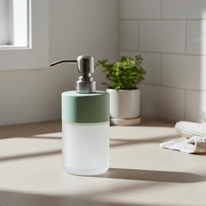 Op de afbeelding: Een zeepdispenser van matglas met een mintgroene bovenkant en een roestvrijstalen pomp. De dispenser is cilindrisch en staat op een lichtgekleurd aanrecht. Een kleine plant in een pot staat op de achtergrond.