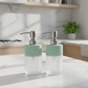 Op de afbeelding: Twee zeepdispensers van matglas met mintgroene bovenkanten en zilveren pompmechanismen. De dispensers zijn cilindrisch en staan op een wit aanrechtblad. De achtergrond omvat een keuken met een plant en houten planken.