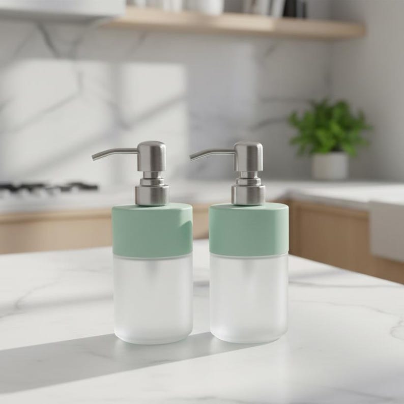 Op de afbeelding: Twee zeepdispensers van matglas met mintgroene bovenkanten en zilveren pompmechanismen. De dispensers zijn cilindrisch en staan op een wit aanrechtblad. De achtergrond omvat een keuken met een plant en houten planken.