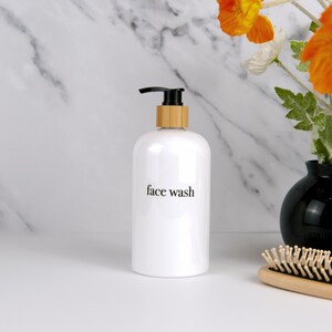 May include: A white face wash bottle with a black pump and a bamboo collar. The words "face wash" are printed in black on the front. The bottle is on a white surface with a marble background and a black vase with orange flowers.